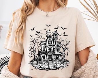 May include: Beige t-shirt featuring a black and white haunted house graphic with bats, bare trees, and jack-o'-lanterns. The design is centered on the shirt and evokes a Halloween theme. The shirt has short sleeves and a crew neck.