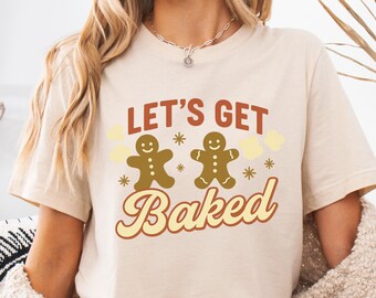 May include: A beige t-shirt featuring the phrase "LET'S GET Baked" in red and yellow. The design includes two gingerbread men and star shapes. A casual, festive top.