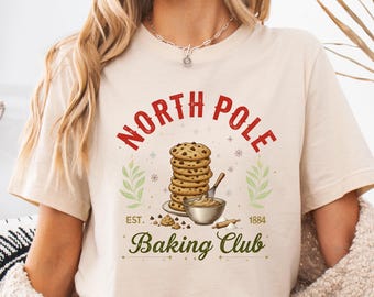 May include: A cream-coloured t-shirt with a graphic that reads "North Pole Baking Club" in red and green. The design includes a stack of chocolate chip cookies, a bowl of batter, and the year 1884.
