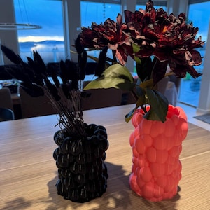 May include: Two 3D printed vases, one black and one pink, with different textured surfaces. The pink vase holds a bouquet of dark red flowers, while the black vase holds a bunch of dried black grasses.