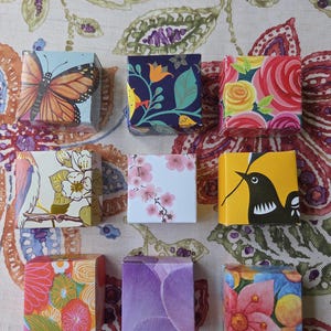 May include: Nine small square boxes arranged in a grid, each featuring a distinct pattern. Designs include a monarch butterfly, floral motifs with roses and cherry blossoms, a black bird on a yellow background, and abstract purple petals.