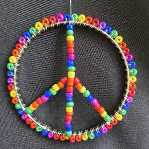 May include: A colorful peace sign ornament made of rainbow-colored beads strung on silver wire. The beads are arranged in a circular shape with three downward-pointing lines, creating the peace symbol.