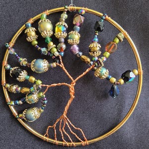 May include: A handcrafted tree of life ornament. The copper wire tree is adorned with colorful beads in shades of blue, green, gold, and purple. The tree is set within a gold-colored circular frame, creating a decorative piece.