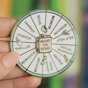 May include: A round enamel pin with a book theme. The pin features a wheel design with book genres like "Fantasy," "Horror," and "Romance." The center has a book with the text "WHAT TO READ." The pin is held by a person.