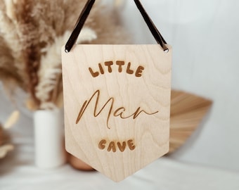 May include: Wooden sign with the text "Little Man Cave" in a brown font. The sign is hanging from a brown string.