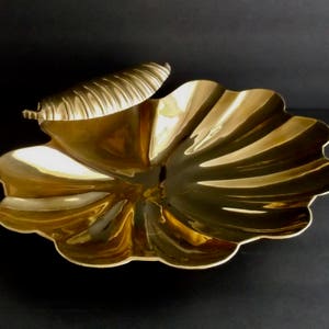 May include: A gold-colored brass shell-shaped dish with a scalloped edge. The dish has a smaller shell-shaped handle on the top.
