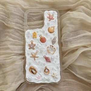 May include: A white phone case decorated with seashells, pearls, and starfish. The case has a clear back and a white front with a textured surface.