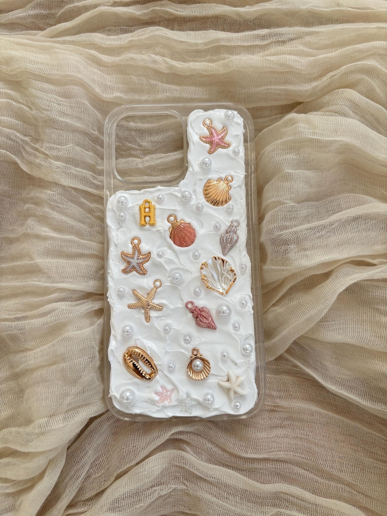 May include: A white phone case decorated with seashells, pearls, and starfish. The case has a clear back and a white front with a textured surface.