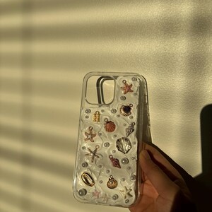 May include: A clear phone case with a white background and various sea-themed charms, including seashells, starfish, and pearls.