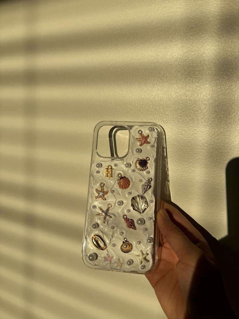 May include: A clear phone case with a white background and various sea-themed charms, including seashells, starfish, and pearls.
