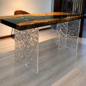 Op de afbeelding: Een rechthoekige tafel met een houten blad met natuurlijke randen en een donkere harsinleg die een vijverscène met koikarpers en waterlelies toont. De tafel wordt ondersteund door twee transparante, getextureerde acrylpoten met een golvend oppervlak.