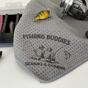 Può includere: Un asciugamano da pesca grigio con la scritta "FISHING BUDDIES" e un'illustrazione di due persone che pescano. Sono visibili anche un mulinello e una scatola per esche con esche. Un'esca gialla e nera è sull'asciugamano.