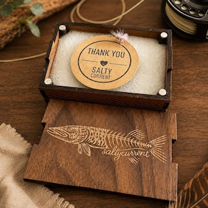 Pode incluir: Uma caixa de madeira escura com tampa deslizante com um esqueleto de peixe gravado a laser e o texto "saltycurrent". Dentro, uma etiqueta redonda bege diz "THANK YOU" com um ícone de coração e "SALTY CURRENT", junto a uma mosca de pesca.