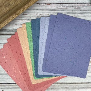 May include: A set of ten sheets of handmade paper in a rainbow of colors, including pink, orange, yellow, green, blue, and purple. The paper has a speckled texture and is perfect for crafting or scrapbooking.