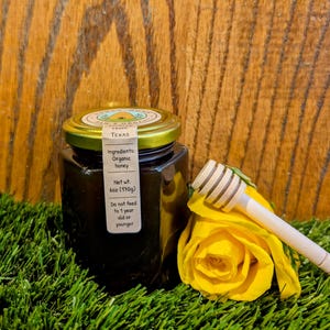 May include: A square glass jar of organic honey from Texas, with a gold lid and a label detailing ingredients and net weight (6 oz / 170g). A wooden honey dipper rests near a yellow rose on a bed of green grass.