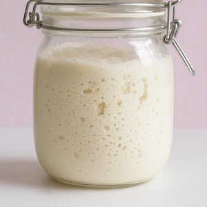 May include: A glass jar with a metal lid and a metal clasp. The jar is filled with a white, bubbly liquid. The liquid is a sourdough starter.