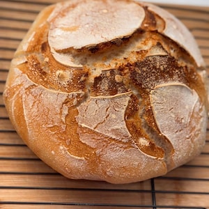 May include: A freshly baked sourdough loaf with a golden brown crust and a slightly cracked surface. The bread is sitting on a wire rack.