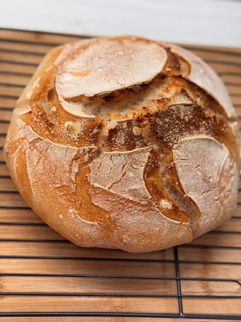 May include: A freshly baked sourdough loaf with a golden brown crust and a slightly cracked surface. The bread is sitting on a wire rack.