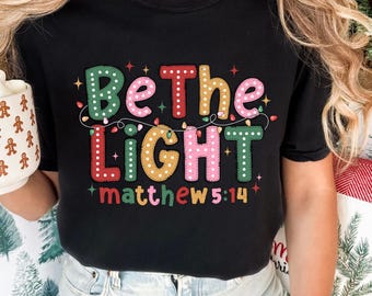 Peut inclure: T-shirt noir avec un graphique coloré qui dit "Soyez la lumière" avec des lumières de Noël et l'Écriture "Matthieu 5:14".