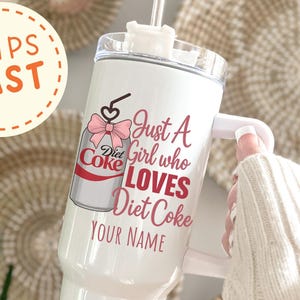 May include: White tumbler with a handle and a straw, featuring a Diet Coke design with a pink bow and the text "Just A Girl who LOVES Diet Coke YOUR NAME". The tumbler is held by a person.