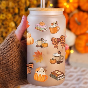 May include: A frosted glass tumbler with a bamboo lid and a stainless steel straw. The tumbler is decorated with fall-themed stickers, including pumpkins, coffee mugs, and a bow. The background features pumpkins and fall foliage.