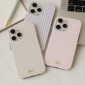 May include: Three phone cases with a gingham pattern in cream, lavender, and pink. Each case has a gold monogram and is next to a laptop, notebook, and pen. The notebook is open to a blank page with the month of April visible.