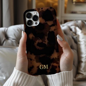 May include: A brown and black tortoiseshell phone case with a gold monogram of the letters "GM". The case has a glossy finish and a camera cutout. The phone case is held in a person's hands.