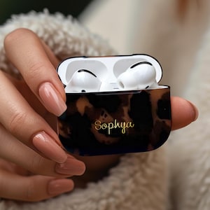 May include: A black and brown leopard print AirPods case with the name "Sophya" in gold script. The case is open, revealing white earbuds. The background is blurred, with a soft, neutral color.