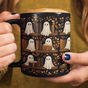 May include: Black ceramic mug featuring a whimsical design of ghost characters on bookshelves. The mug has a black handle and is filled with a coffee drink. The design includes autumn leaves and books, creating a cozy, seasonal theme.