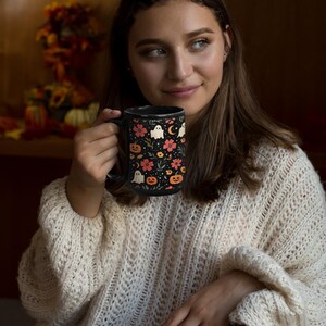 May include: Black ceramic mug with a Halloween-themed design. The mug features a pattern of ghosts, pumpkins, flowers, and a crescent moon on a black background. The handle is also black.