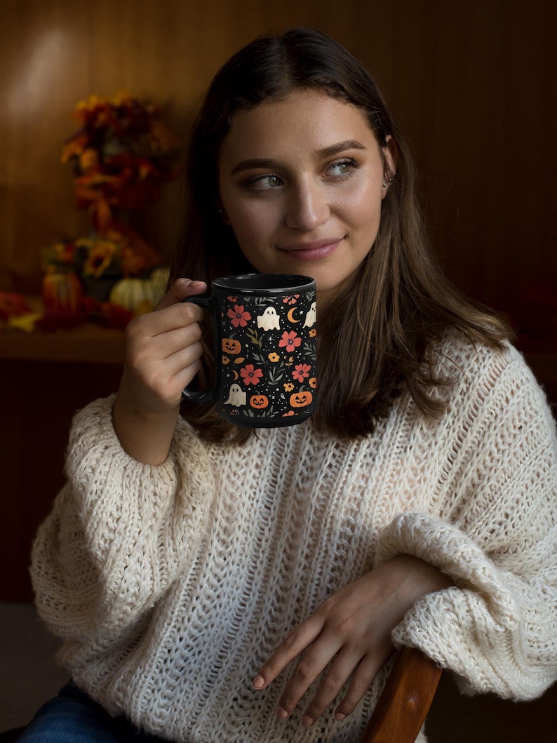 May include: Black ceramic mug with a Halloween-themed design. The mug features a pattern of ghosts, pumpkins, flowers, and a crescent moon on a black background. The handle is also black.