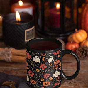 May include: A black ceramic mug filled with a dark liquid, featuring a Halloween-themed pattern of ghosts, pumpkins, flowers, and moons. The mug is set on a wooden surface with a gold spoon and surrounded by candles and pumpkins.