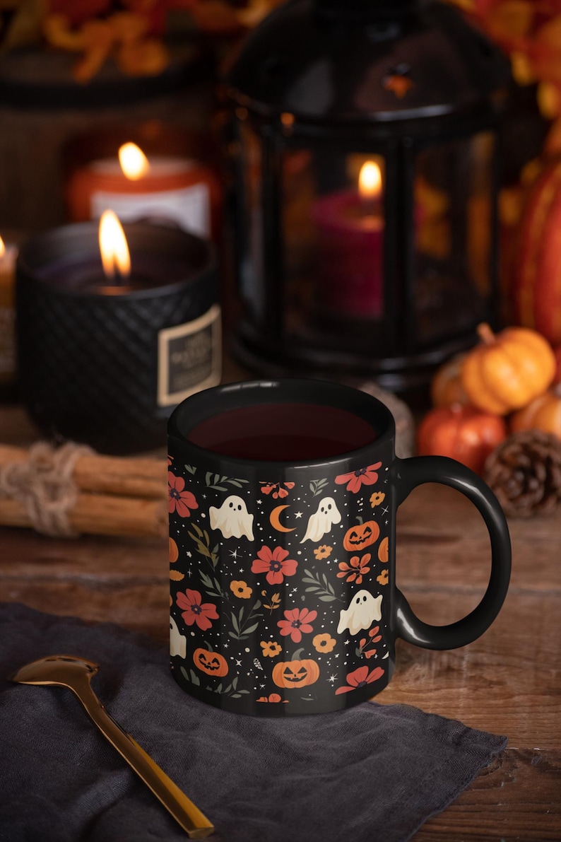 May include: A black ceramic mug filled with a dark liquid, featuring a Halloween-themed pattern of ghosts, pumpkins, flowers, and moons. The mug is set on a wooden surface with a gold spoon and surrounded by candles and pumpkins.