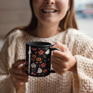 May include: Black ceramic mug featuring a Halloween-themed pattern. The design includes orange pumpkins, white ghosts, and pink flowers on a black background. The mug has a black handle and interior.