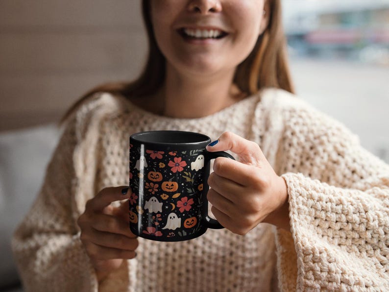 May include: Black ceramic mug featuring a Halloween-themed pattern. The design includes orange pumpkins, white ghosts, and pink flowers on a black background. The mug has a black handle and interior.