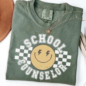 May include: Olive green t-shirt with the words "SCHOOL COUNSELOR" in white block letters around a yellow smiley face with lightning bolt eyes. The shirt has a checkerboard pattern and a Comfort Colors tag.