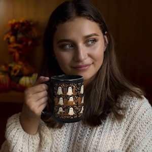 May include: Black ceramic mug featuring a repeating pattern of white ghosts on bookshelves with autumn leaves. The mug has a large handle and is held by a person wearing a cream-colored sweater. The background is blurred, with fall foliage visible.