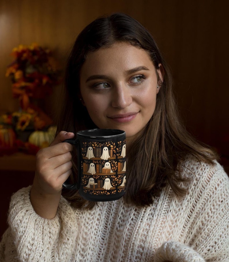 May include: Black ceramic mug featuring a repeating pattern of white ghosts on bookshelves with autumn leaves. The mug has a large handle and is held by a person wearing a cream-colored sweater. The background is blurred, with fall foliage visible.