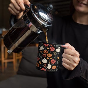 May include: A black coffee mug with a Halloween-themed pattern of white ghosts, orange pumpkins, and pink flowers. Coffee is being poured from a French press into the mug. The mug has a black handle.