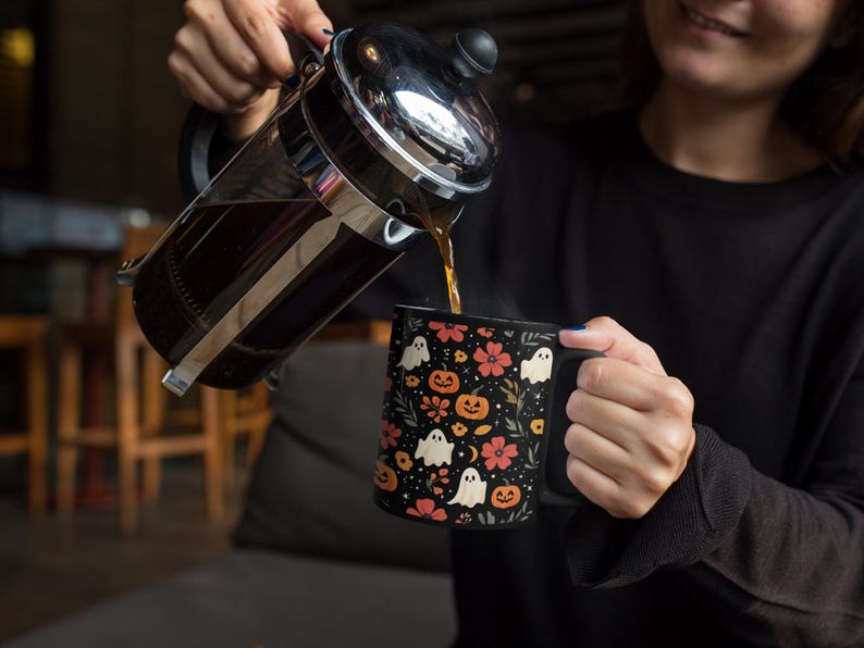May include: A black coffee mug with a Halloween-themed pattern of white ghosts, orange pumpkins, and pink flowers. Coffee is being poured from a French press into the mug. The mug has a black handle.