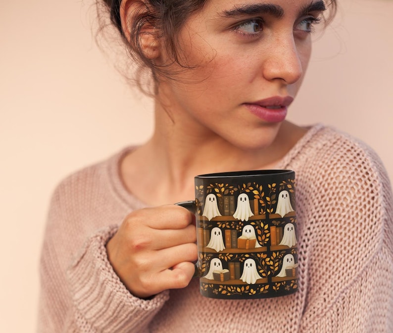 May include: Black ceramic mug featuring a repeating pattern of white ghost figures on bookshelves with books and autumn leaves. The mug has a black handle and a black interior. The design is set against a black background.