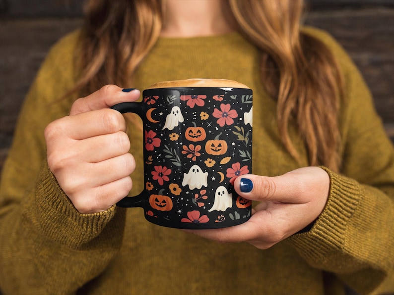 May include: Black ceramic mug with a Halloween-themed pattern. The design features orange pumpkins, white ghosts, red flowers, and crescent moons on a black background. The mug is filled with a creamy beverage.