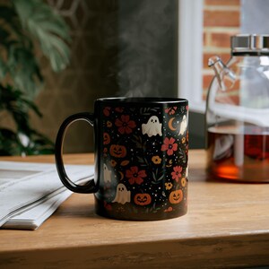 May include: Black ceramic mug with a Halloween-themed design. The mug features a pattern of white ghosts, orange pumpkins, red flowers, and a crescent moon on a black background. Steam rises from the mug, suggesting a hot beverage.