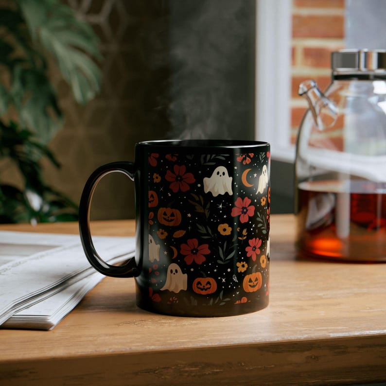 May include: Black ceramic mug with a Halloween-themed design. The mug features a pattern of white ghosts, orange pumpkins, red flowers, and a crescent moon on a black background. Steam rises from the mug, suggesting a hot beverage.