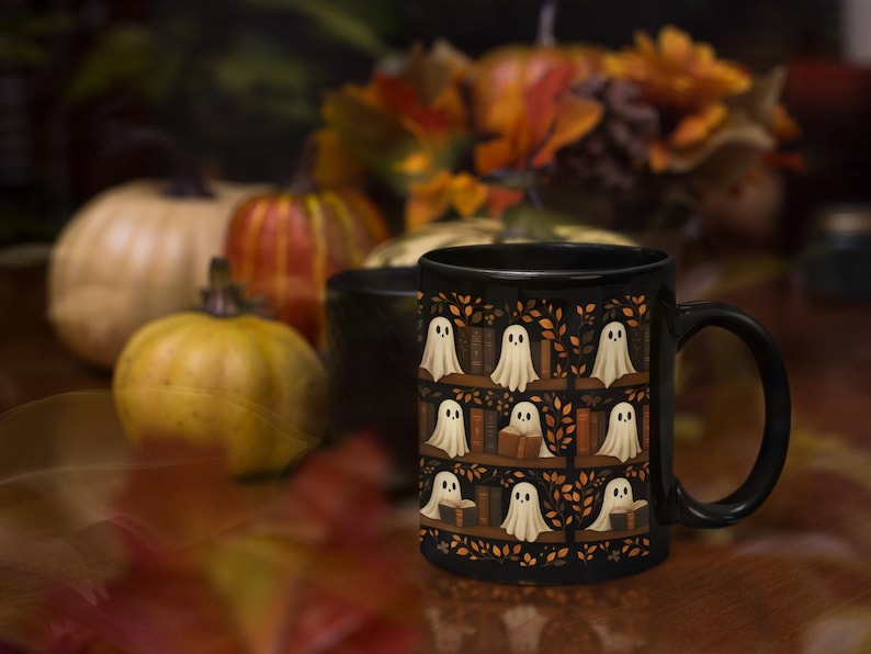 May include: Black ceramic mug featuring a repeating pattern of white ghost figures on bookshelves, with autumn leaves and pumpkins in the background. The mug has a black handle and is likely for hot beverages.
