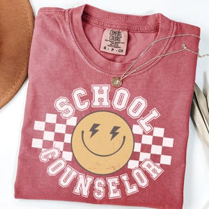May include: A red Comfort Colors t-shirt with the words "SCHOOL COUNSELOR" in white block letters surrounding a yellow smiley face with lightning bolt eyes. A silver necklace with a small pendant rests on the shirt.