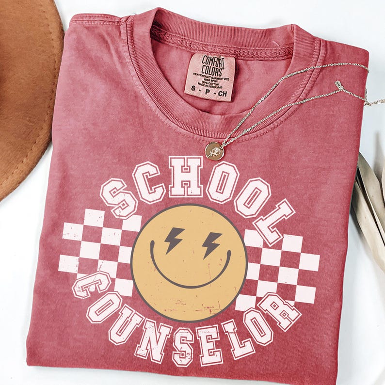 May include: A red Comfort Colors t-shirt with the words "SCHOOL COUNSELOR" in white block letters surrounding a yellow smiley face with lightning bolt eyes. A silver necklace with a small pendant rests on the shirt.