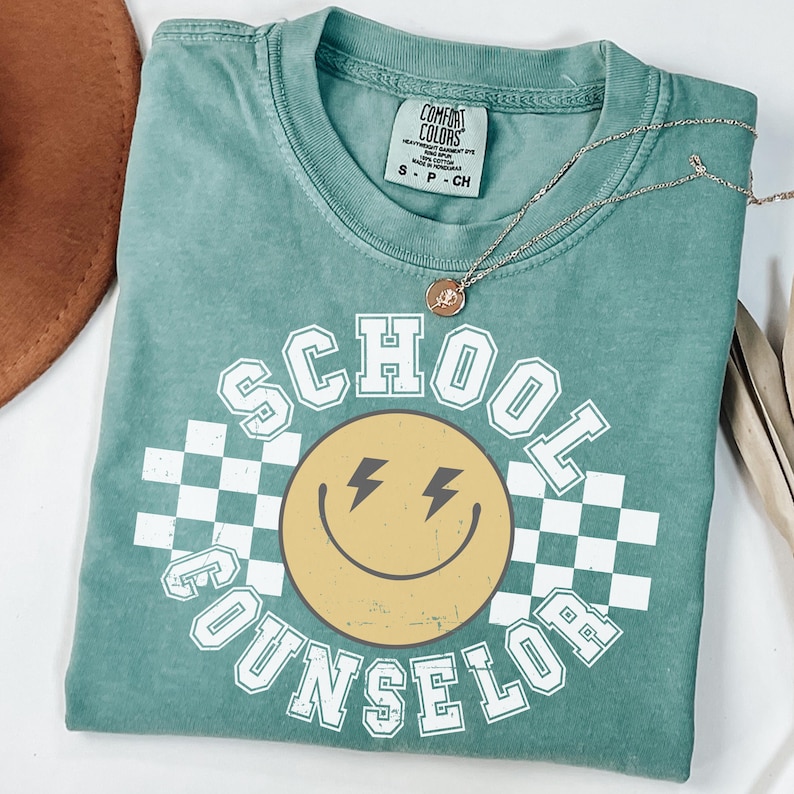May include: A teal Comfort Colors t-shirt with the words "SCHOOL COUNSELOR" in white block letters. A yellow smiley face with lightning bolt eyes is in the center, set against a white and black checkered background. A gold necklace is draped across the shirt.