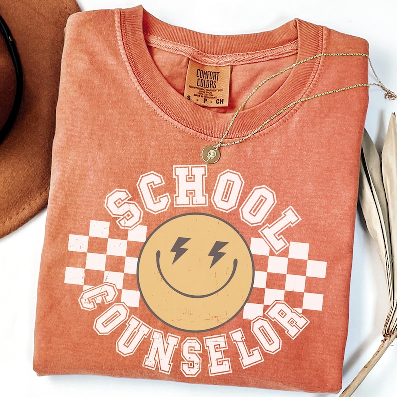 May include: An orange Comfort Colors t-shirt with the words "SCHOOL COUNSELOR" surrounding a smiling face with lightning bolt eyes. A gold necklace and a brown hat are also visible.