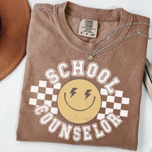 May include: A brown t-shirt with the words "SCHOOL COUNSELOR" in white block letters. A yellow smiley face with lightning bolt eyes is in the center, set against a white and black checkered background. A silver necklace with a small pendant rests on the shirt.